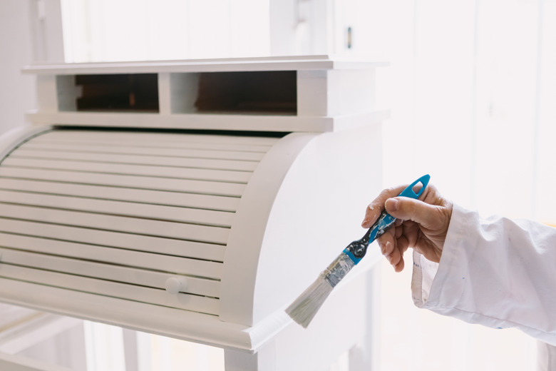 Detail of hand of person in white coat painting a piece of wooden furniture white with a brush.