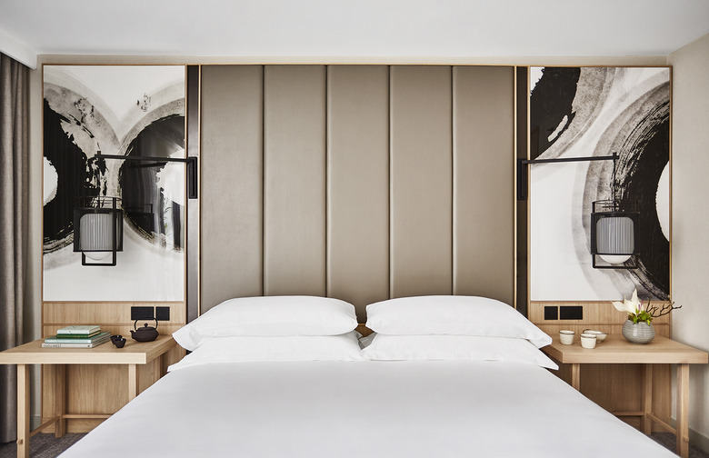 King size bed with white beddings and pillows with ceiling-high fabric-paneled headboard with wood side tables and paintings above