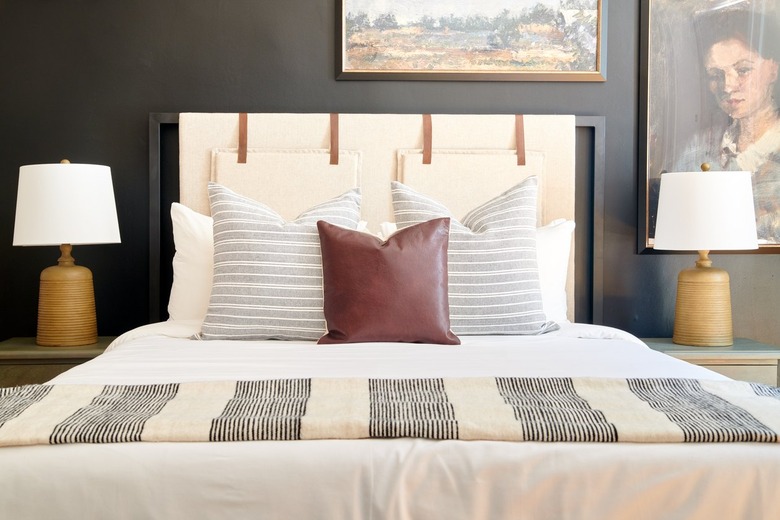 Modern bedroom with fabric headboard, matching lamps and nightstands, paintings on wall, dark gray wall.