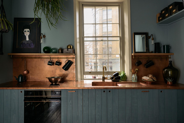 Rustic colors in kitchen with muted teal cabinets, copper counters and backsplash, open shelves, cream trim on window.