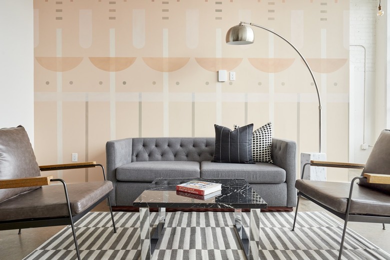 Modern living room with gray couch, large metal floor lamp, mirror coffee table, leather armchairs, striped rug, wallpaper.