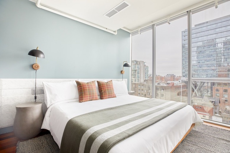 Modern bedroom with window to ceiling windows, gray nightstand, white bedding, light green blanket, accent pillows with orange and green, jute rug.