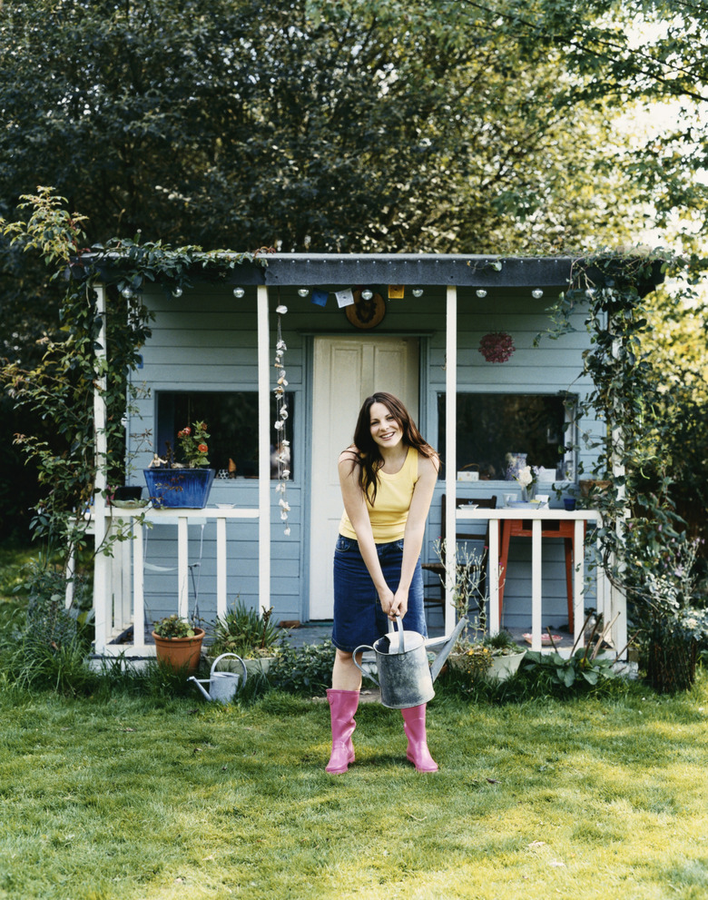 Woman Stood in Front of a Small Wooden Bungalow, Holding a Watering Can