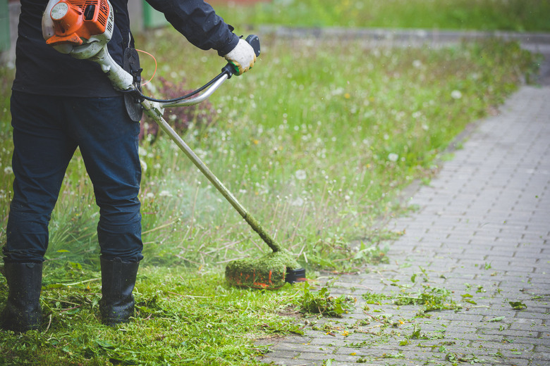 Worker in dark protective clothing with a gas mower in his hands, mowing grass in front of the house. A man mows grass with dandelions on a rainy spring day. Trimmer in the hands of a man.