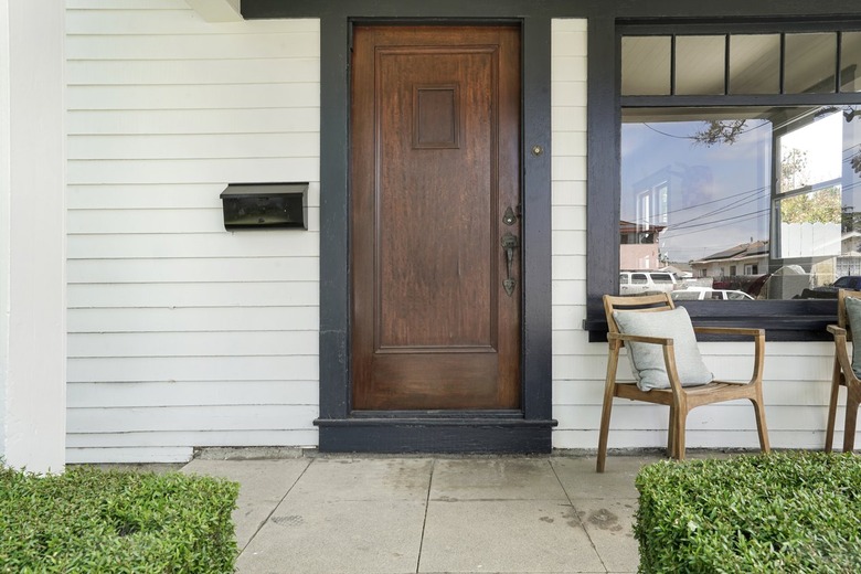 A wooden front door on a white house with a chair on the front porch