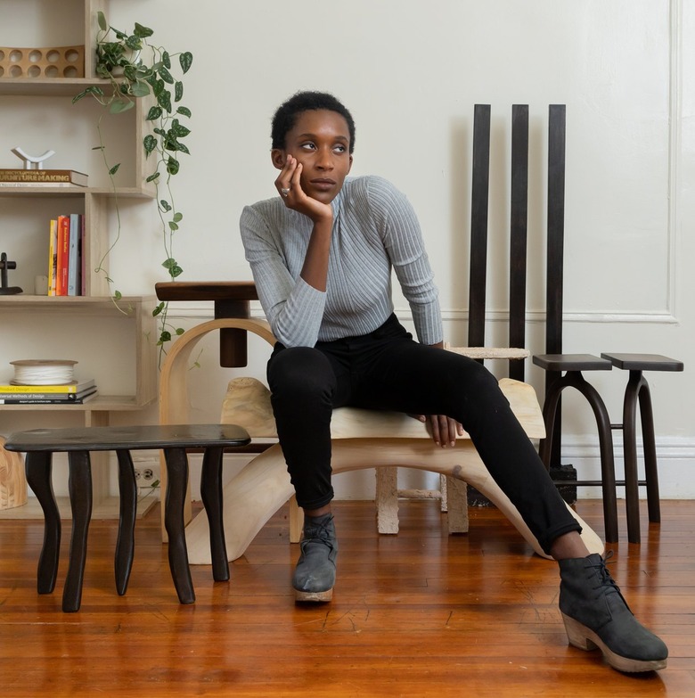 Furniture designer Nifemi Ogunro sitting on a piece of wood furniture, wearing black pants, black suede cloglike boots, and a long light gray T-shirt.