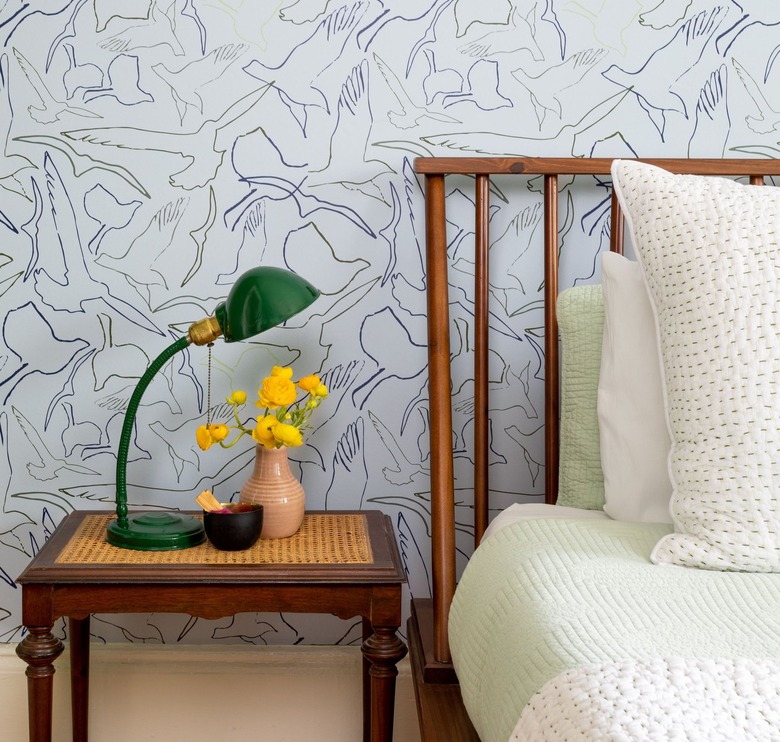 Light blue wallpaper with birds behind a bed with a wooden frame and a wooden side table.