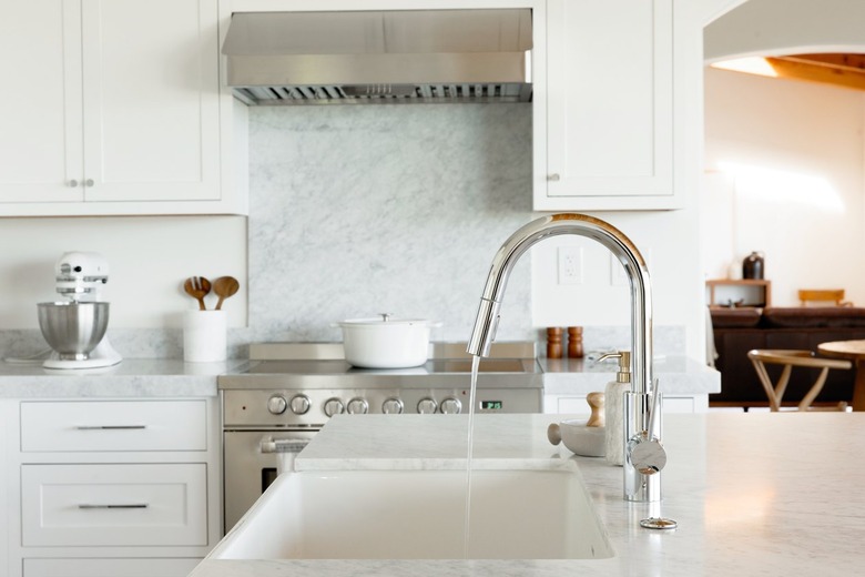 In a kitchen, a sink in a marble countertop, part of a kitchen island. The chrome faucet is running water. In the background, there