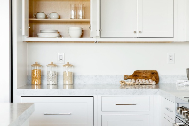 Kitchen counter. The countertop itself is marble, and the cabinets above and below are white. The top left cabinet is open, revealing a stacks of white plates, white bowls, two white mugs, and two clear glasses. On the counter, three glass food storage containers, a cutting board, and a string of garlic.