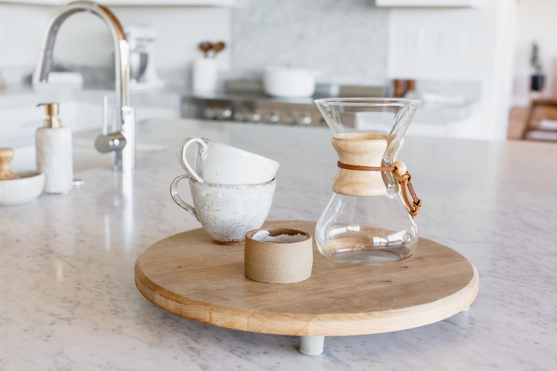 On a marble countertop, two stacked ceramic mugs, a Chemex, and a candle in a ceramic holder on a small round wooden board. In the background, a chrome faucet and a marbled soap dispenser.