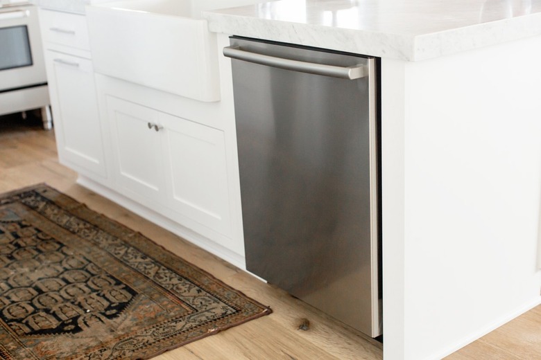 Stainless steel dishwasher in a white kitchen island. The marble countertop extends over the dishwasher. On the floor, a small rug.