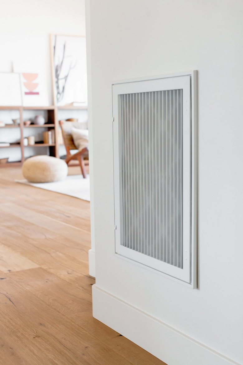 A white air vent on a white wall. Living room partially visible in the distance.