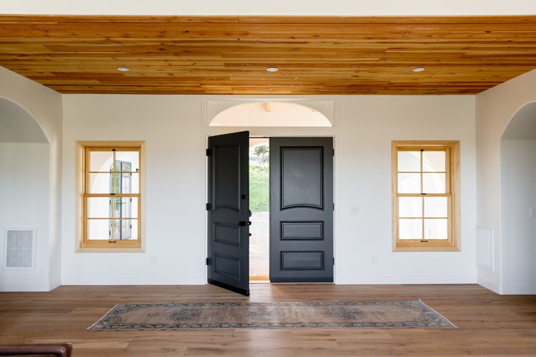 An entry hall of a Spanish-style home with double dark wood doors, one of which is open, on a white wall. Two windows with light wood frames on either side of the door. The floor and ceiling are both made of wood. There