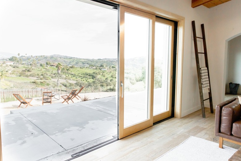 Glass sliding doors with a light wood frame are half-open, leading from a living room to an outdoor patio. The walls of the living room are white and there