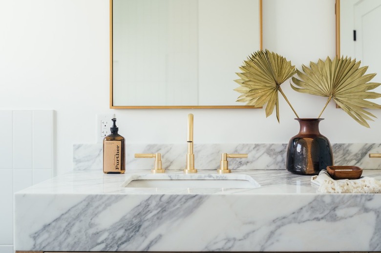 Bathroom sink with white and gray countertop, brass or gold faucet, and mirror. Vase with dried plant.