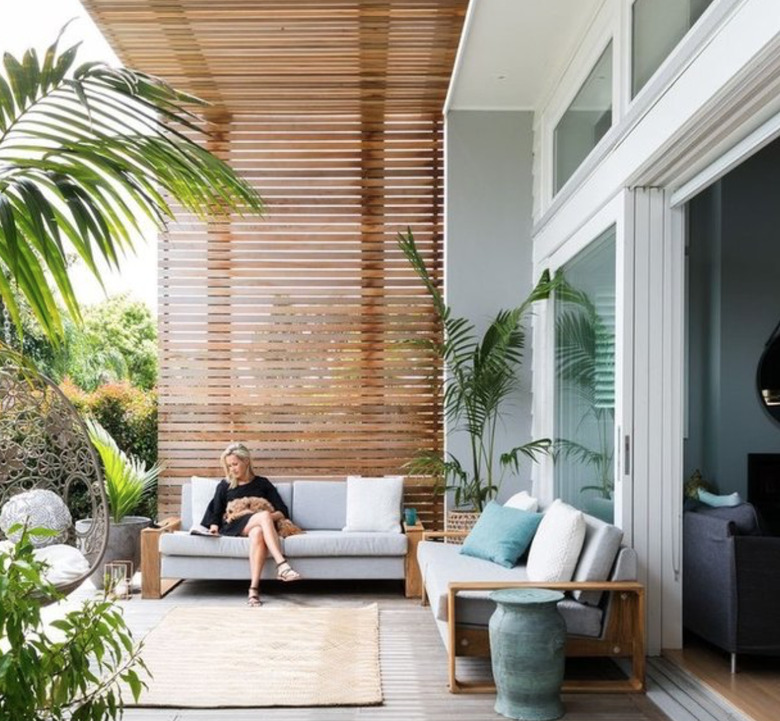 Modern pergola with two outdoor couches, rug, palms, woman seated.