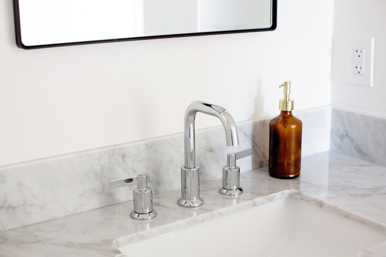 A granite or marble counter with a silver faucet sink. A brown glass dispenser bottle with a gold top is beside and a mirror above.