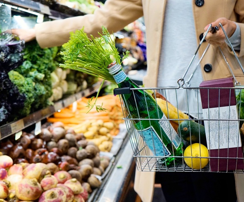 Woman in tan pea coat placing produce into a grocery basket.