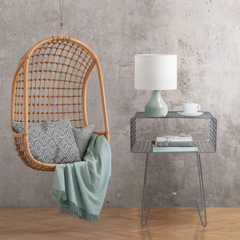 Image of a metal side table next to a chair swing. The chair swing is accessorized with a grey throw pillow and a teal blanket throw. The nightstand is accessorized with a teal and white lamp, a teacup, and books.