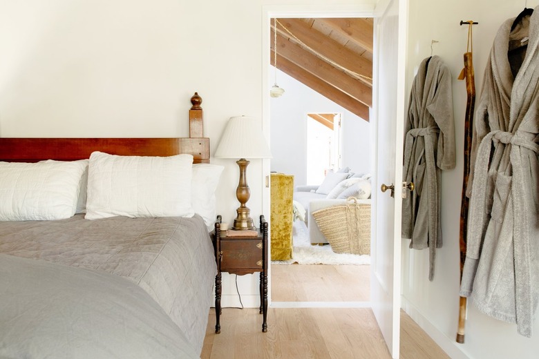 Bedroom with wood headboard, vintage night table and lamp. Wall with hanging bathrobes and a wood walking stick.