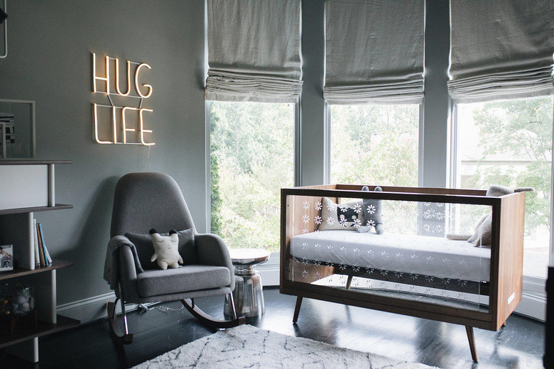 Gray baby nursery idea with crib in front of bay windows with Roman shades and neon sign hanging on wall above rocking chair