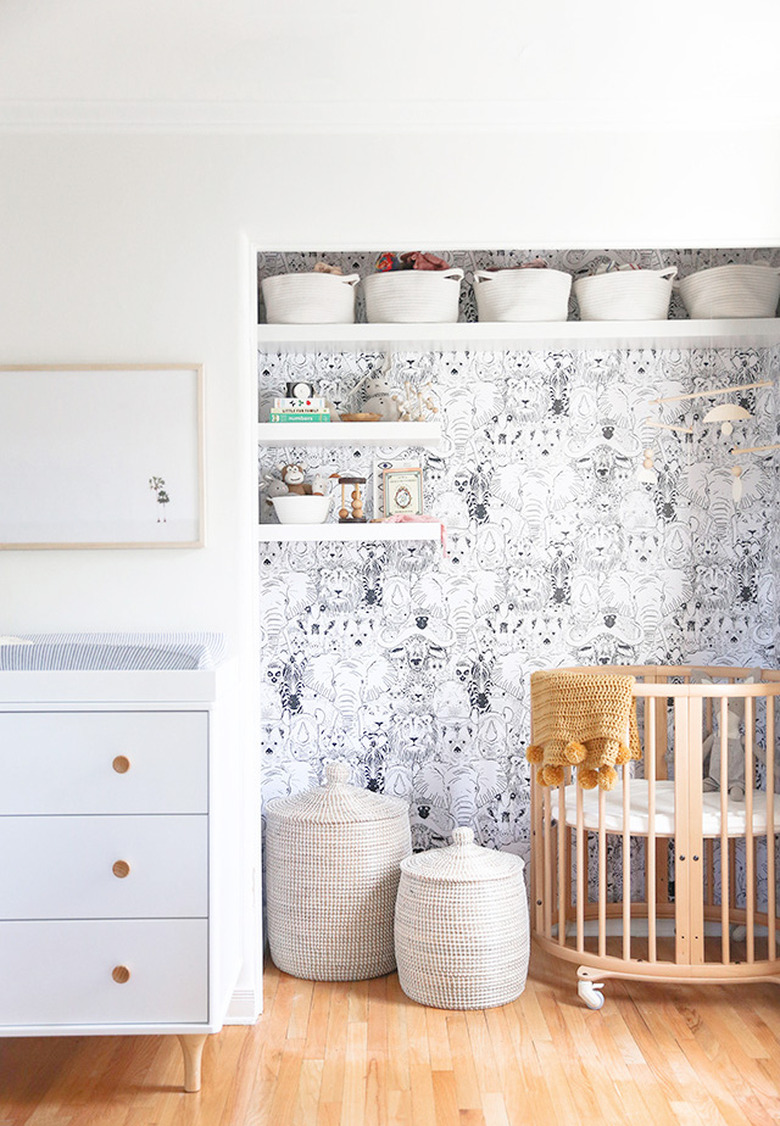 Nursery with wallpaper in nook, round crib, baskets, shelves, white dresser.