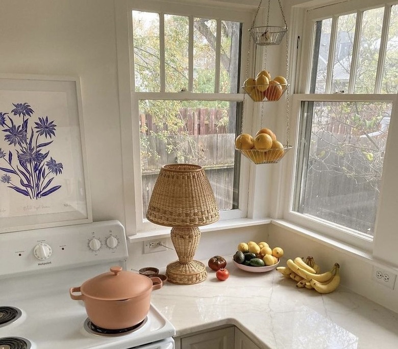 A pink Perfect Pot rests on a white stovetop with a blue and white floral painting, a wicker lamp, a hanging fruit basket, and a bowl of fruit on the countertop.