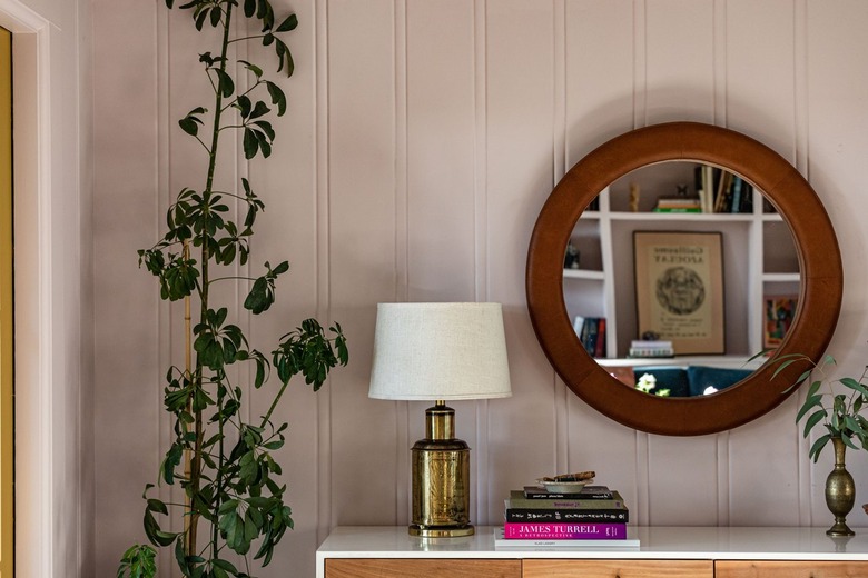 Round wood mirror over a credenza with a gold lamp and books. A plant is beside them.