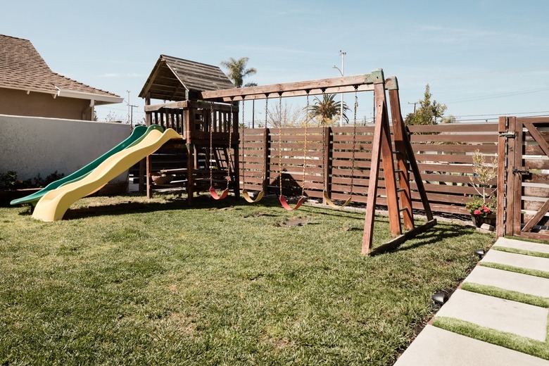 A wooden swing set on top of a patch of grass; a wooden horizontal fence and square concrete pavers