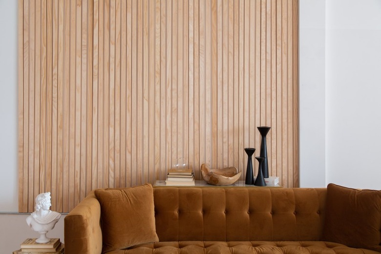 Brown tufted velvet couch, with a wood beam accent wall, white bust sculpture, and black Modernist style vases.