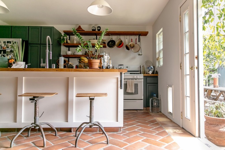 Kitchen with terra-cotta floor, white kitchen island, industrial wood stools, a white bell pendant light, green cabinets and wood shelves.