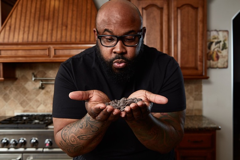 Philip Ashley Rix, a Black man in a black T-shirt wearing black glasses, is holding chocolate chips in his hands and pursing his lips. It almost looks as though he is blowing on the chocolate.