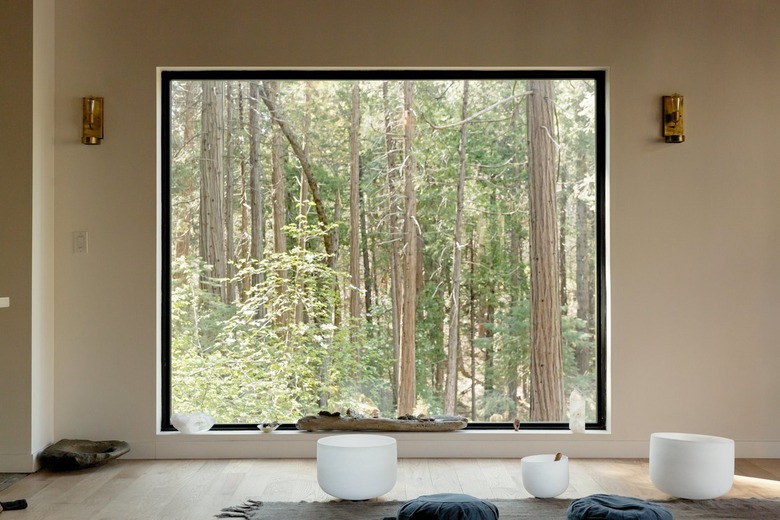 A large picture window with a forrest view, and crystals. A neutral rug with blue cushions, and white meditation singing bowls.