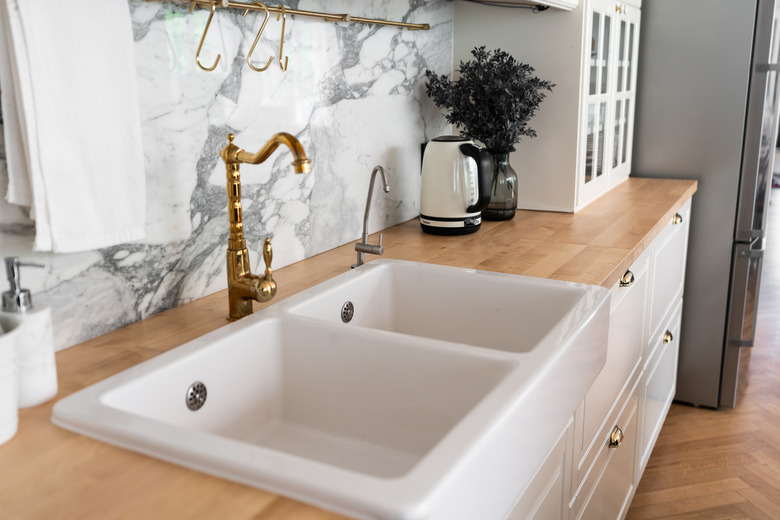 Modern classic kitchen interior with kitchen appliances and white ceramic sink with gold mirror faucet on wood top with marble wall in the background / kitchen interior