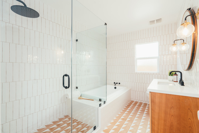 glass walk-in shower, orange patterned floor tile, white vertical subway wall tile, wood vanity with white countertop, round mirror, bathtub