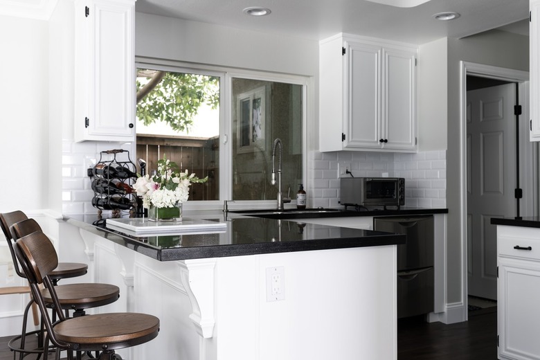 A kitchen with white cabinets, black counters, white tile backsplash, and wood bar stools. A wine rack and flowers, on a counter.