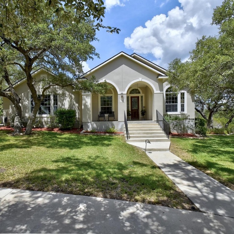 Exterior of an off-white home in San Antonio, Texas. There is a grassy area in front of the stairs along with large trees and a blue sky.