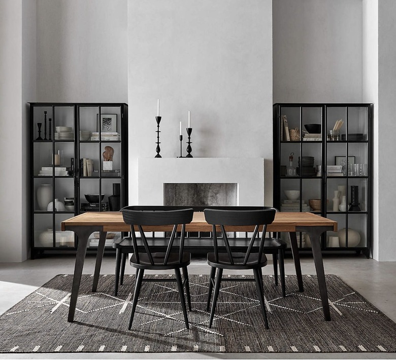 Image of a wooden table with black legs surrounded by four chairs. There's a fireplace behind the table and decorative black shelving with books and accessories on either side of the fireplace.