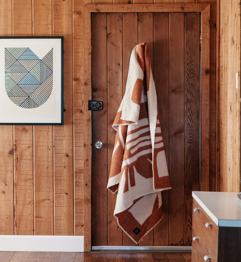 Throw blanket hangs on back of door in wood paneled room, with wood dresser and art print on wall.