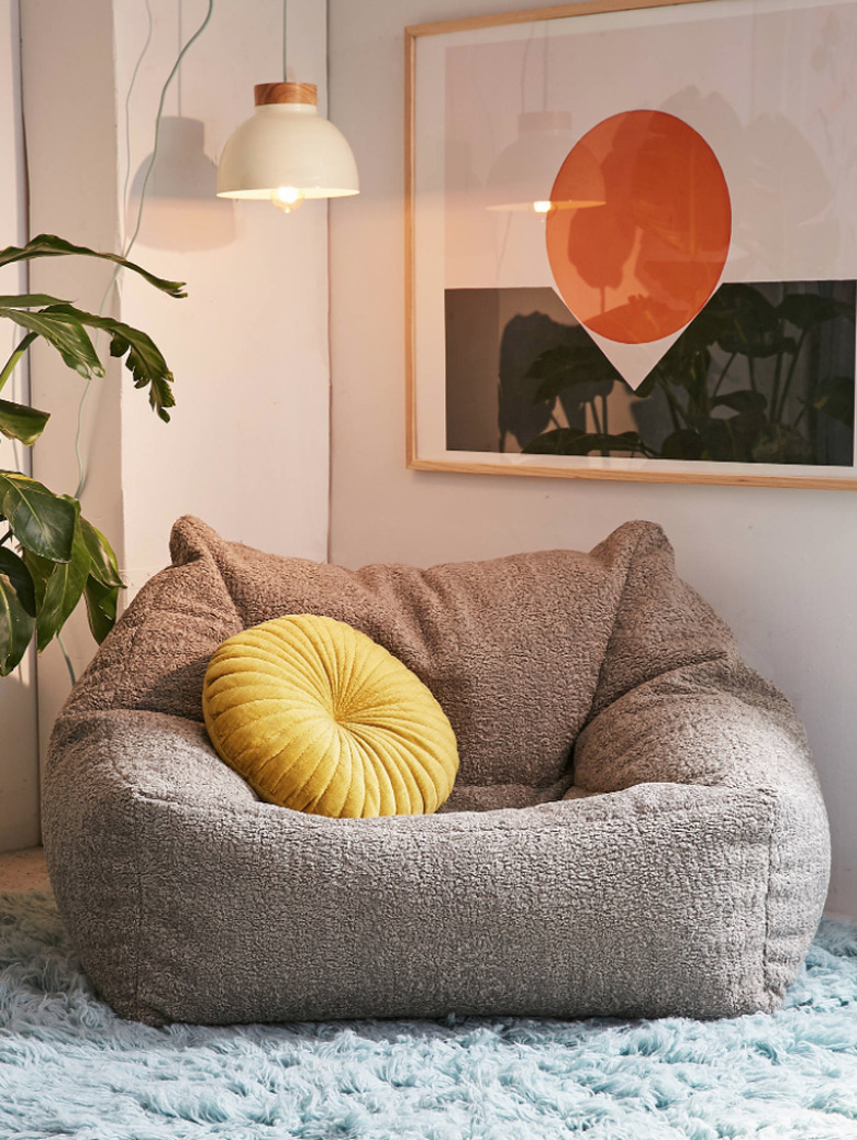 Grey faux sheepskin lounge chair, yellow pillow, light blue shag rub, white pendant lamp, plant, art with orange circle and black band.