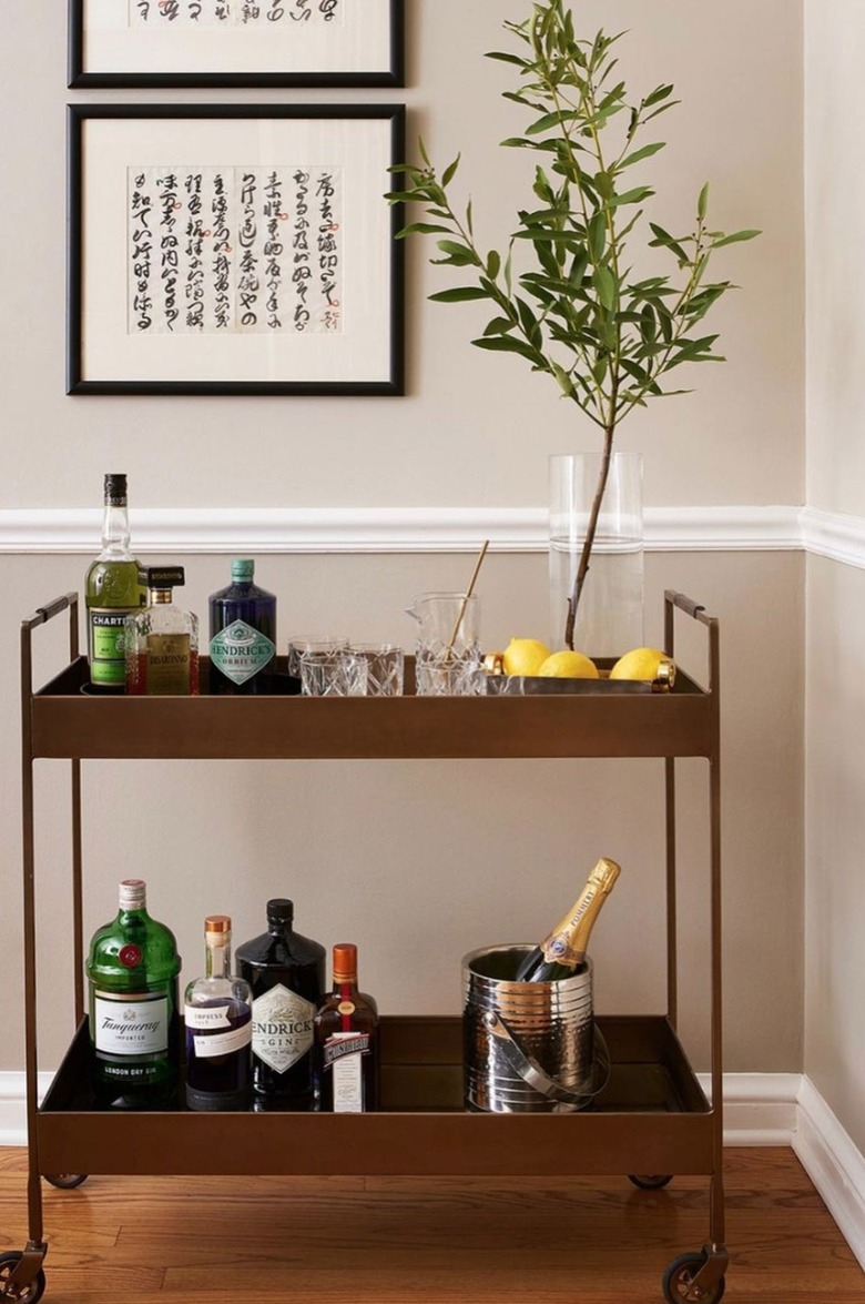 Dark wood and metal bar cart with branch in vase, bottles, glassware and artwork.