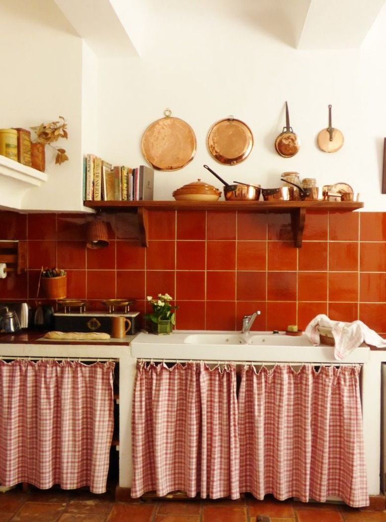 Kitchen with bright orange backsplash, copper pots, open shelving and patterned cabinet curtains.