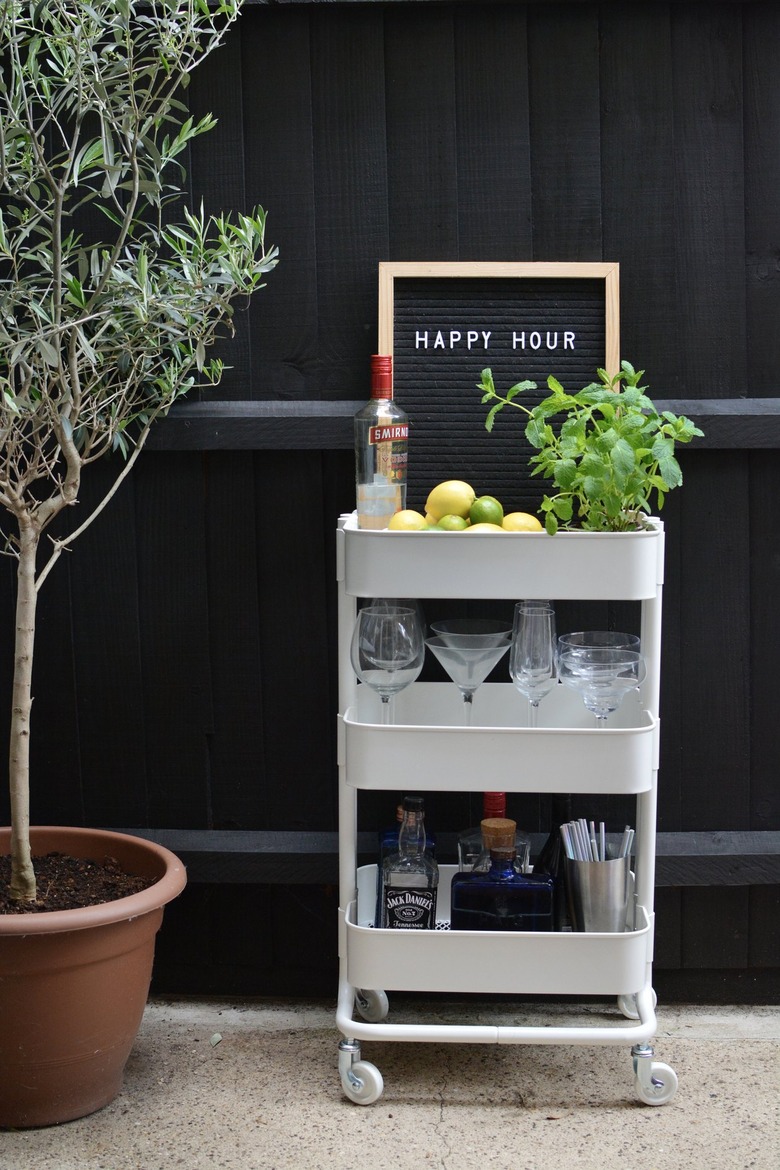 White bar cart with lemons, limes, plant, glasses, liquor bottles next to tree