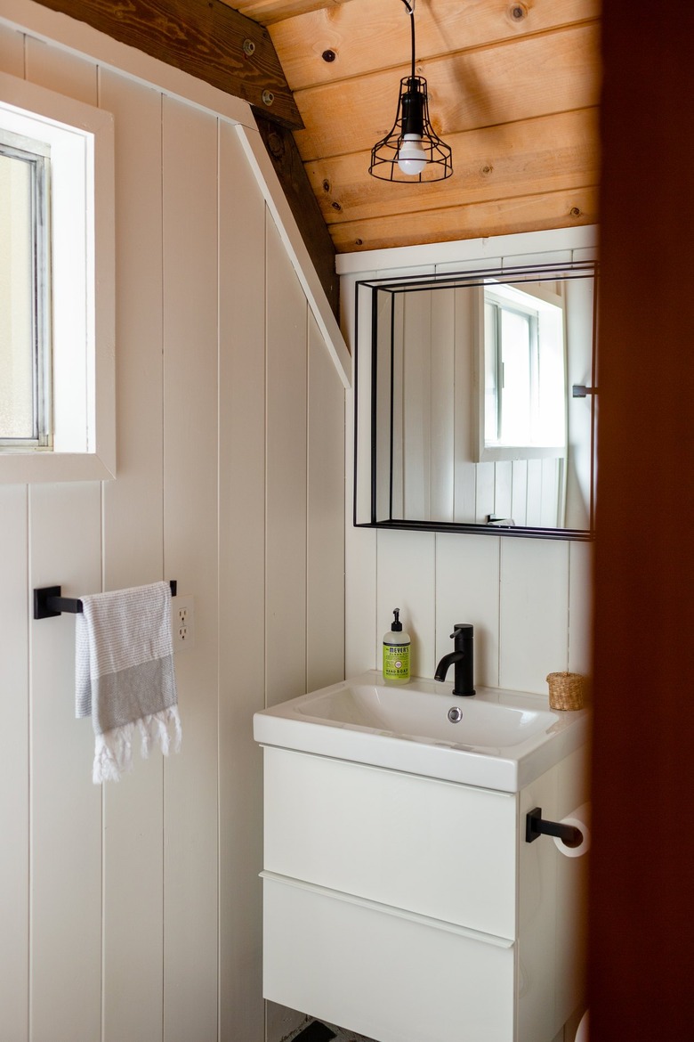 White-walled bathroom with wood ceiling with mirror, small white sink, black faucet, and white hand towel