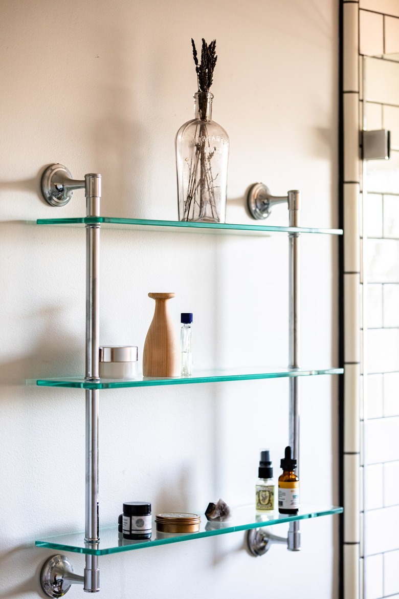 A glass shelving unit attached to a white bathroom wall