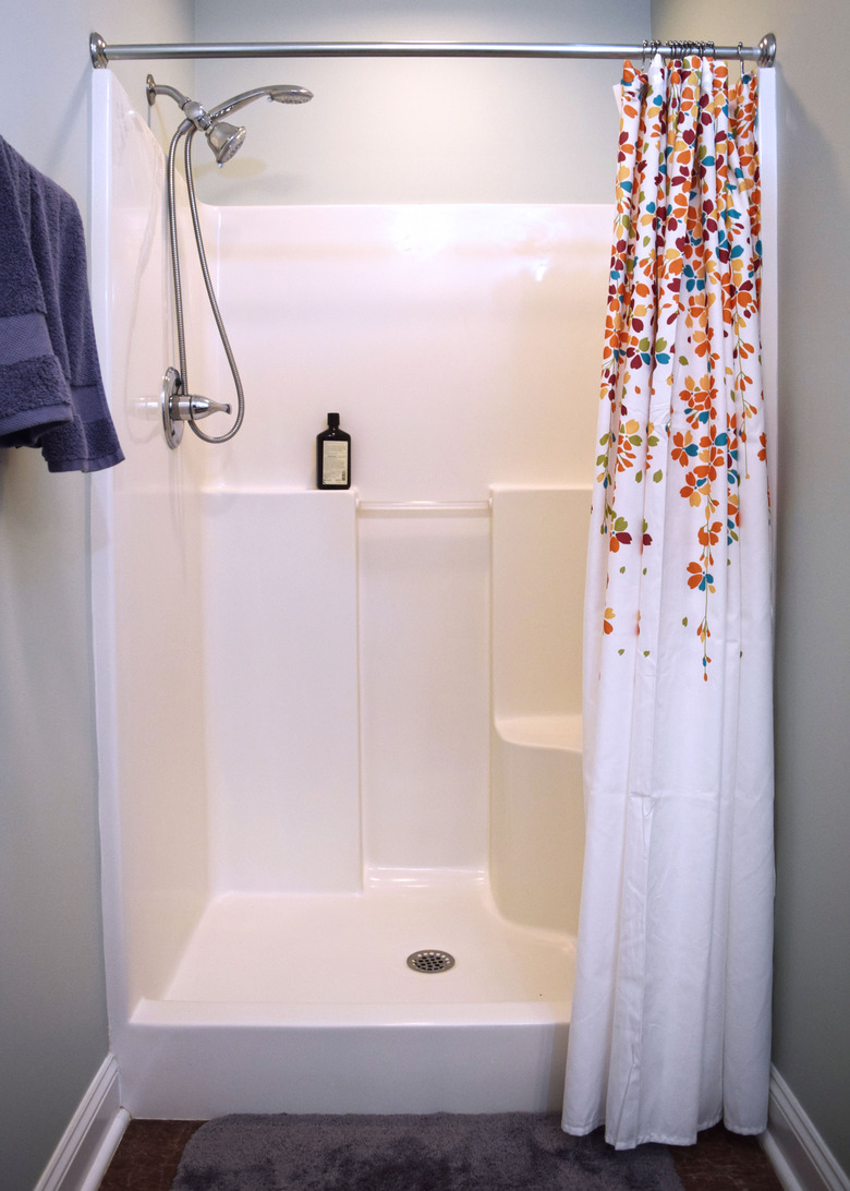 A white walk-in shower with light salt green walls and a weeping floral shower curtain. Purple towels hand on the left wall and a grey bathmat sits on a brown marble tile.