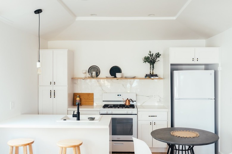 Small kitchen with white cabinets and marble backsplash. Wood open shelving, and one hanging pendant light.