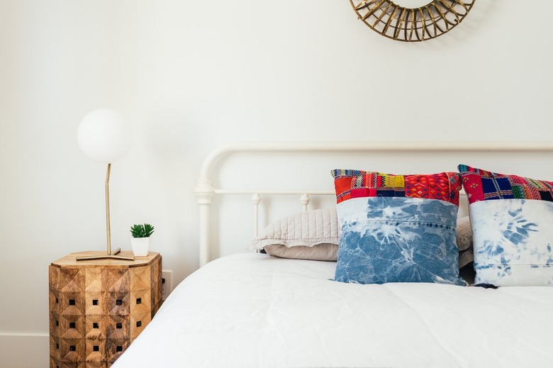 Mirror over a bed with white bedding and multicolored pillows with a wood side table with a globe table lamp and a small plant