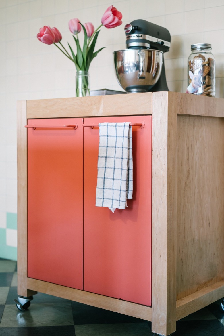 A rollable wood cart with pink cabinet doors. A vase of tulips, mixer, and a jar are on top.