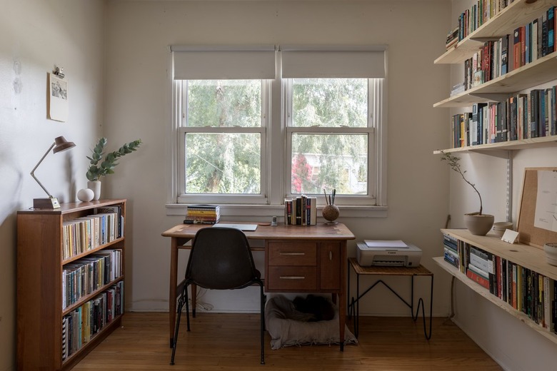 Wood office desk surrounded by bookshelves and shelves of books. Houseplants decorate the space. A printer is on a small table.